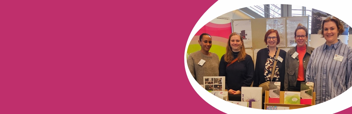 Gruppe von sechs Personen steht hinter einem Tisch mit verschiedenen Produkten und Informationsmaterialien vor einer pink-weißen Wand