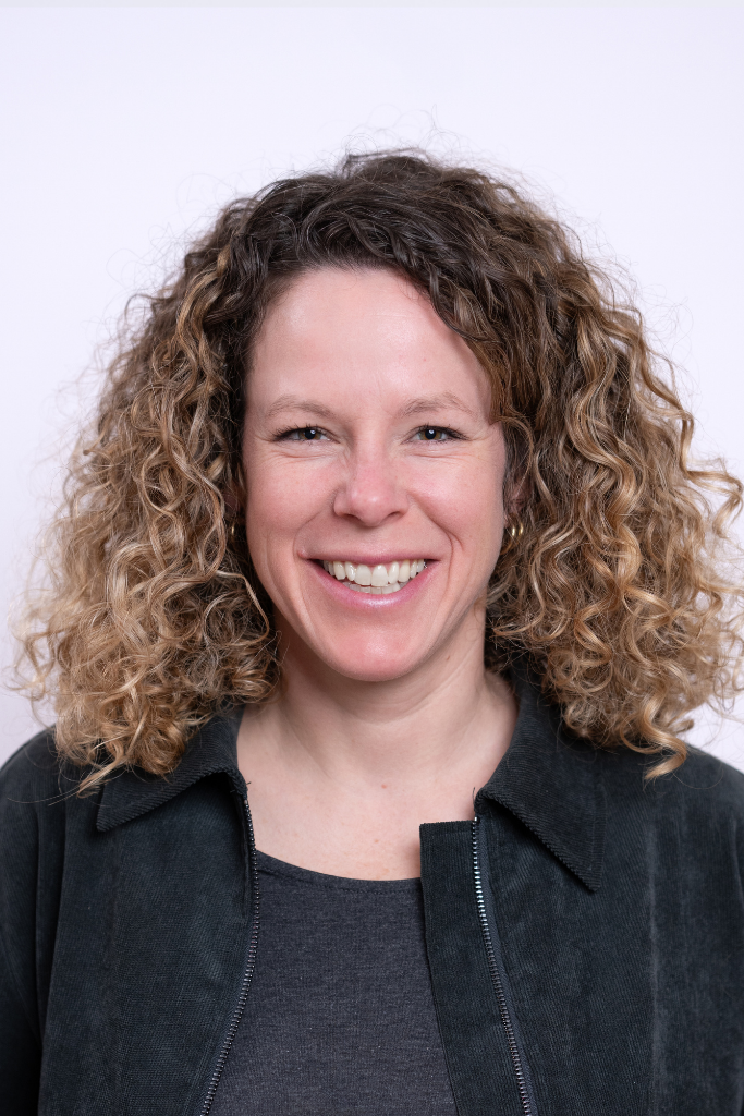 Frau mit mittellangen, lockigen Haaren trägt eine dunkle Jacke über einem dunklen Oberteil vor hellem Hintergrund.