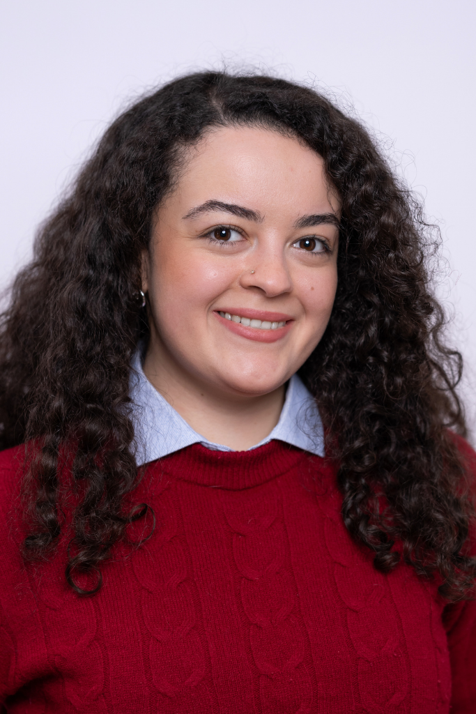 Frau mit langen, lockigen Haaren trägt einen roten Strickpullover über einem Hemd mit Kragen.