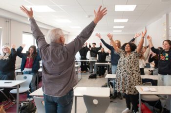 Gruppe von Menschen in einem Klassenraum steht an Tischen und hebt die Arme nach oben