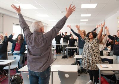 Gruppe von Menschen in einem Klassenraum steht an Tischen und hebt die Arme nach oben