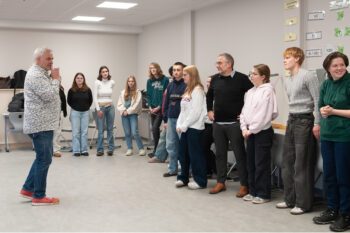 Gruppe von Menschen steht in einem Halbkreis in einem hellen Raum, eine Person spricht vor der Gruppe