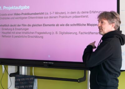 Referent mit Kapuzenpullover zeigt auf einen Bildschirm mit Text zur Projektaufgabe für einen Video-Praktikumsbericht