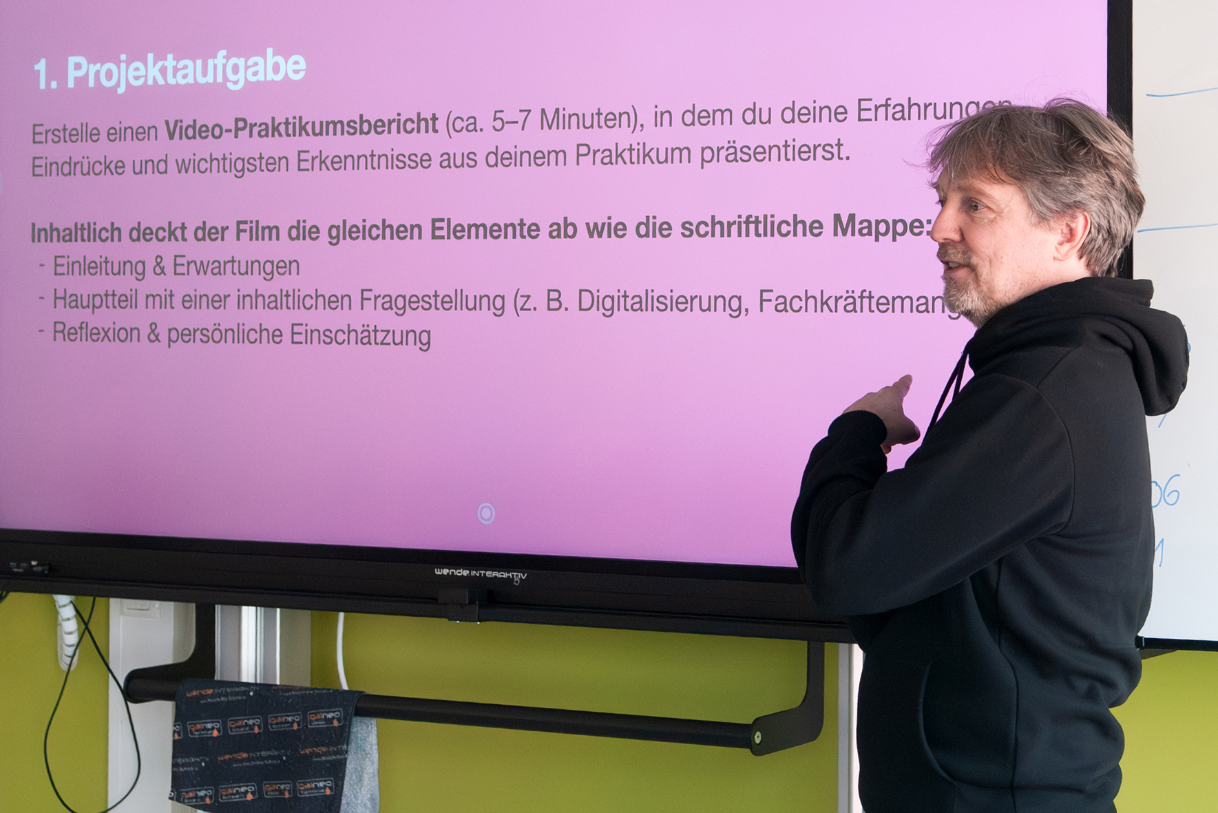 Einführung in den Filmdreh für das Praktikum Referent mit Kapuzenpullover zeigt auf einen Bildschirm mit Text zur Projektaufgabe für einen Video-Praktikumsbericht