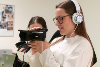 Zwei junge Frauen bei Filmaufnahmen, eine hält eine Kamera, die andere trägt Kopfhörer und hält ein Smartphone