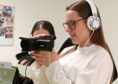 Zwei junge Frauen bei Filmaufnahmen, eine hält eine Kamera, die andere trägt Kopfhörer und hält ein Smartphone