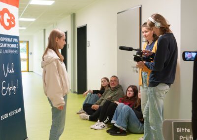 Jugendliche stehen und sitzen in einem Flur, eine Person filmt mit Kamera und Mikrofon, links ein Banner mit dem Schriftzug 'KOOPERATIVE GESAMTSCHULE BAD LAUTERBERG'