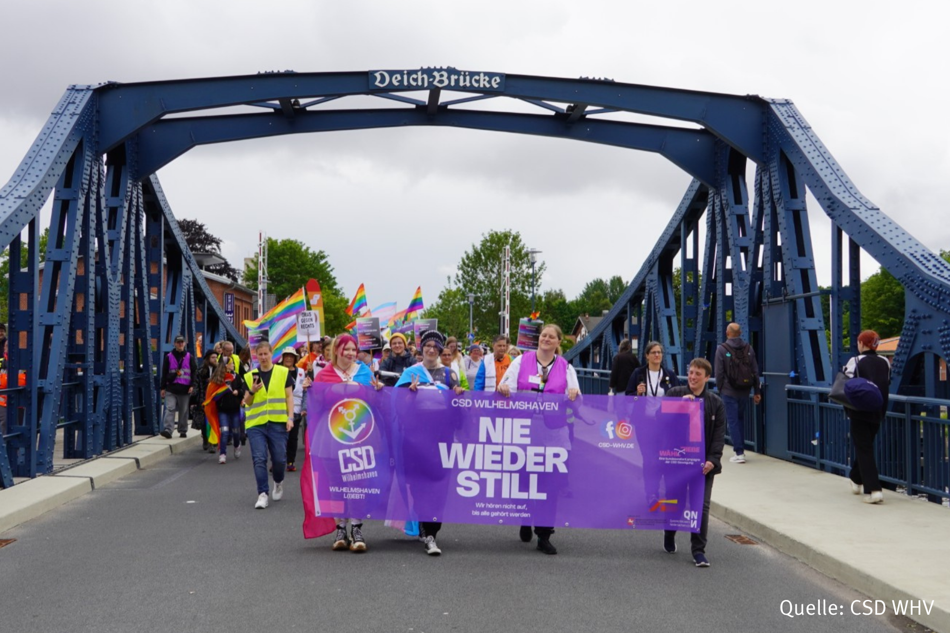 Eine Gruppe von Menschen zieht mit bunten Regenbogenfahnen und einem großen lilafarbenen Banner über eine stählerne Brücke. Das Banner trägt den Schriftzug 'NIE WIEDER STILL'. Im Hintergrund sind Bäume und weitere Teilnehmende zu sehen. Der Himmel ist bewölkt. Am linken und rechten Bildrand sind Hände und Smartphones von Zuschauenden sichtbar.