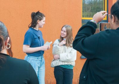 Zwei Mädchen stehen vor einer orangefarbenen Wand, eines hält einen Zettel, das andere ein Mikrofon, im Vordergrund sind zwei Personen von hinten zu sehen