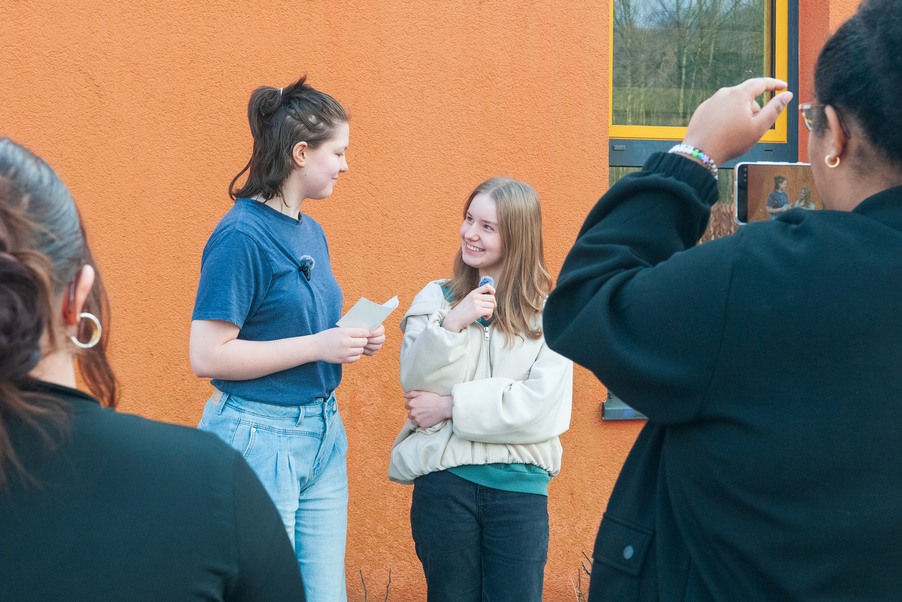 Stärken Interview Zwei Mädchen stehen vor einer orangefarbenen Wand, eines hält einen Zettel, das andere ein Mikrofon, im Vordergrund sind zwei Personen von hinten zu sehen