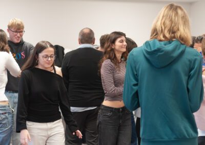 Gruppe von Menschen in einem Raum, einige stehen mit dem Rücken zur Kamera, andere frontal oder seitlich, Kleidung variiert von Pullovern bis zu kurz geschnittenen Oberteilen