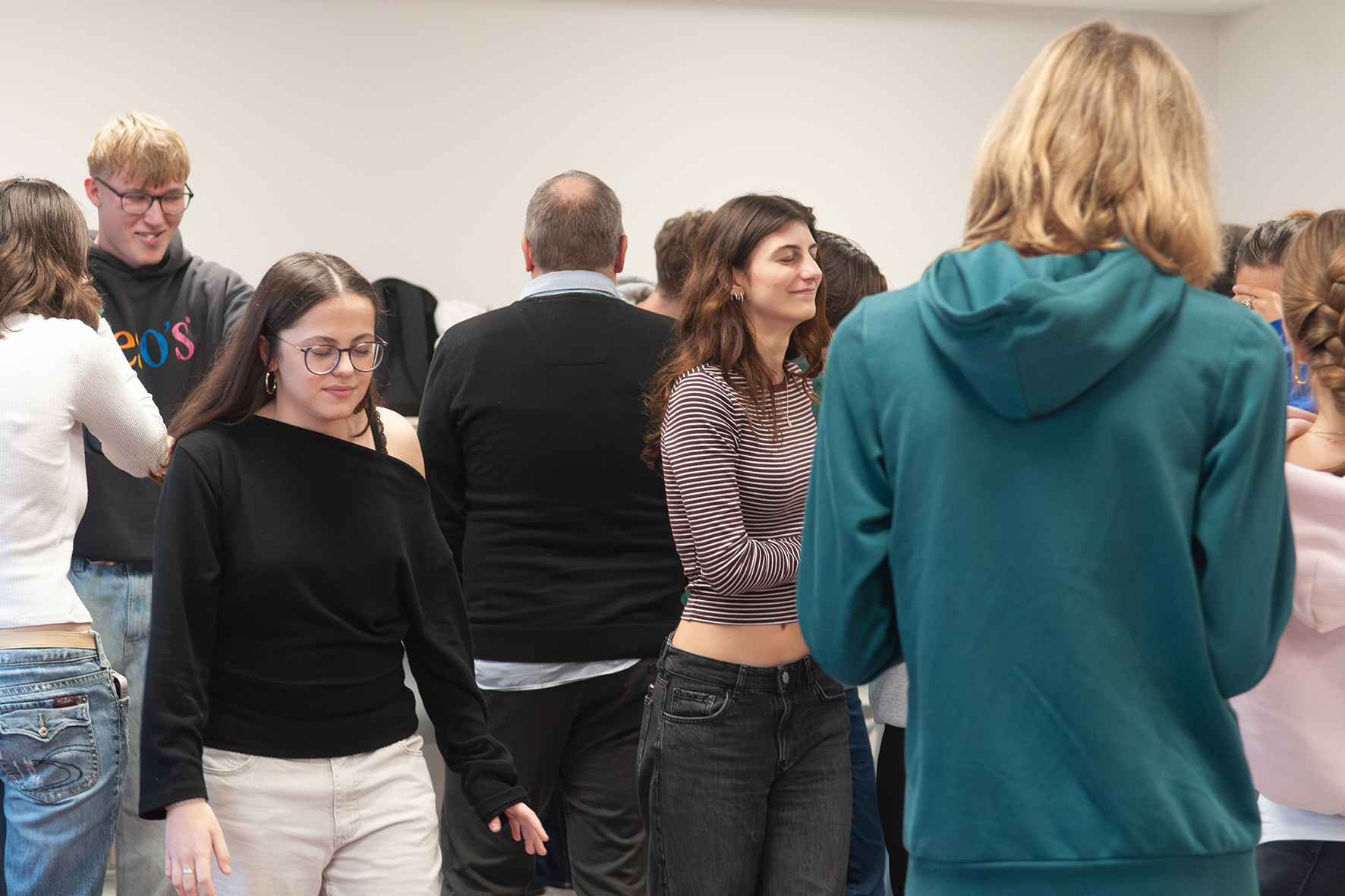 Wahrnehmung im Raum Gruppe von Menschen in einem Raum, einige stehen mit dem Rücken zur Kamera, andere frontal oder seitlich, Kleidung variiert von Pullovern bis zu kurz geschnittenen Oberteilen