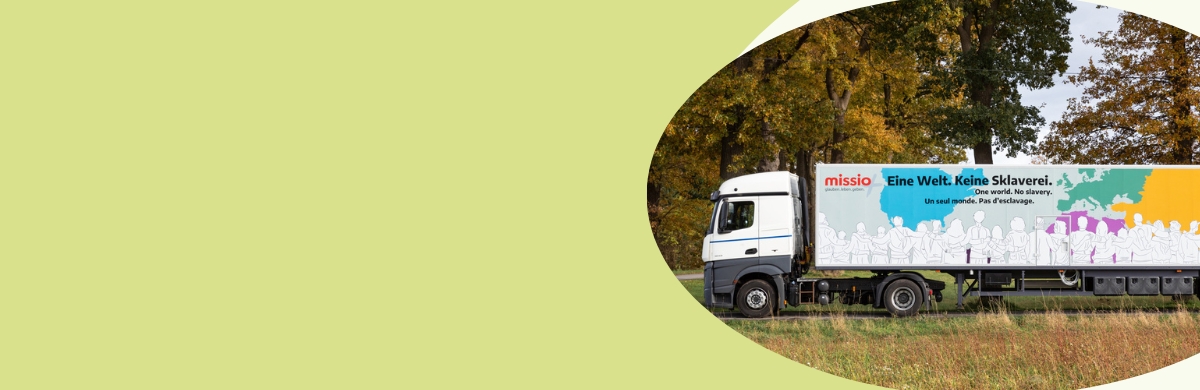Missio Truck Truck LKW mit Aufschrift 'Eine Welt. Keine Sklaverei.' und farbiger Weltkarte auf der Seite, im Hintergrund Bäume