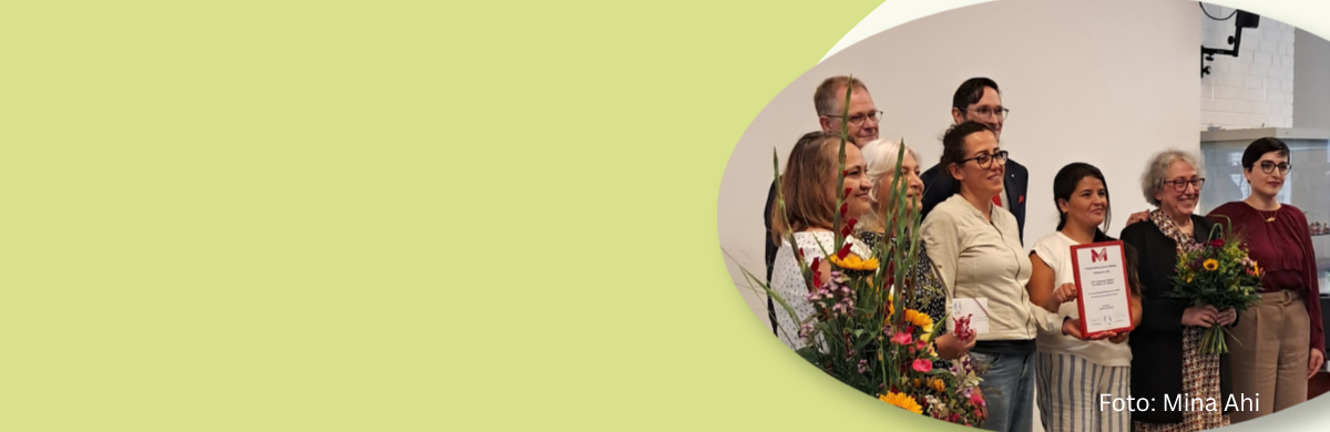 LIVT PREIS SLIDER Gruppe von acht Personen steht in einem Raum, zwei Frauen halten Blumensträuße, eine Frau hält eine Urkunde