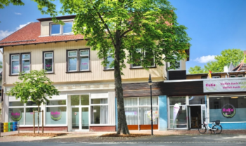 Im Vordergrund steht ein zweistöckiges Gebäude mit hellem Fassadenanstrich und einem roten Satteldach. Große Fenster dominieren die Erdgeschossfront, einige davon sind mit runden, lilafarbenen Aufklebern versehen. Rechts schließt ein flacherer Anbau mit dunkler Verkleidung an, dessen Fenster ebenfalls mit lilafarbenen Kreisen beklebt sind. Über dem Eingang des Anbaus hängt ein weißes Schild mit lilafarbenem und rotem Schriftzug. Ein Fahrrad steht vor dem Gebäude. Ein großer Laubbaum spendet Schatten auf den Gehweg und die Straße.