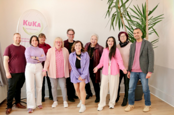 Eine Gruppe von Menschen steht in einem hellen Raum vor einer Wand mit einer großen Pflanze. Die Personen tragen überwiegend farbenfrohe Kleidung in Rosa-, Lila- und Rottönen. Links im Hintergrund befindet sich ein rundes Banner mit dem Schriftzug 'KuKA'. Die Stimmung wirkt freundlich und entspannt, einige Personen lächeln. Der Boden ist hell, die Wand schlicht, und das Licht fällt gleichmäßig auf die Szene.